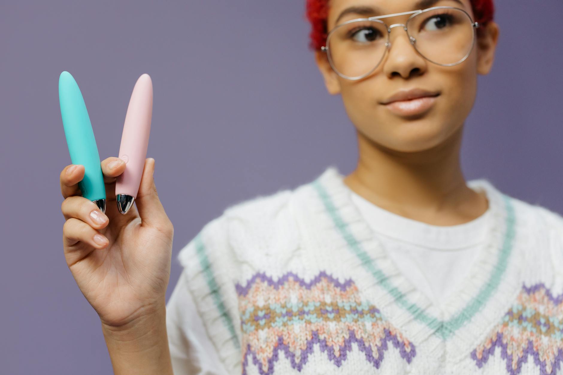 Mulher jovem com óculos segurando vibradores coloridos contra fundo roxo