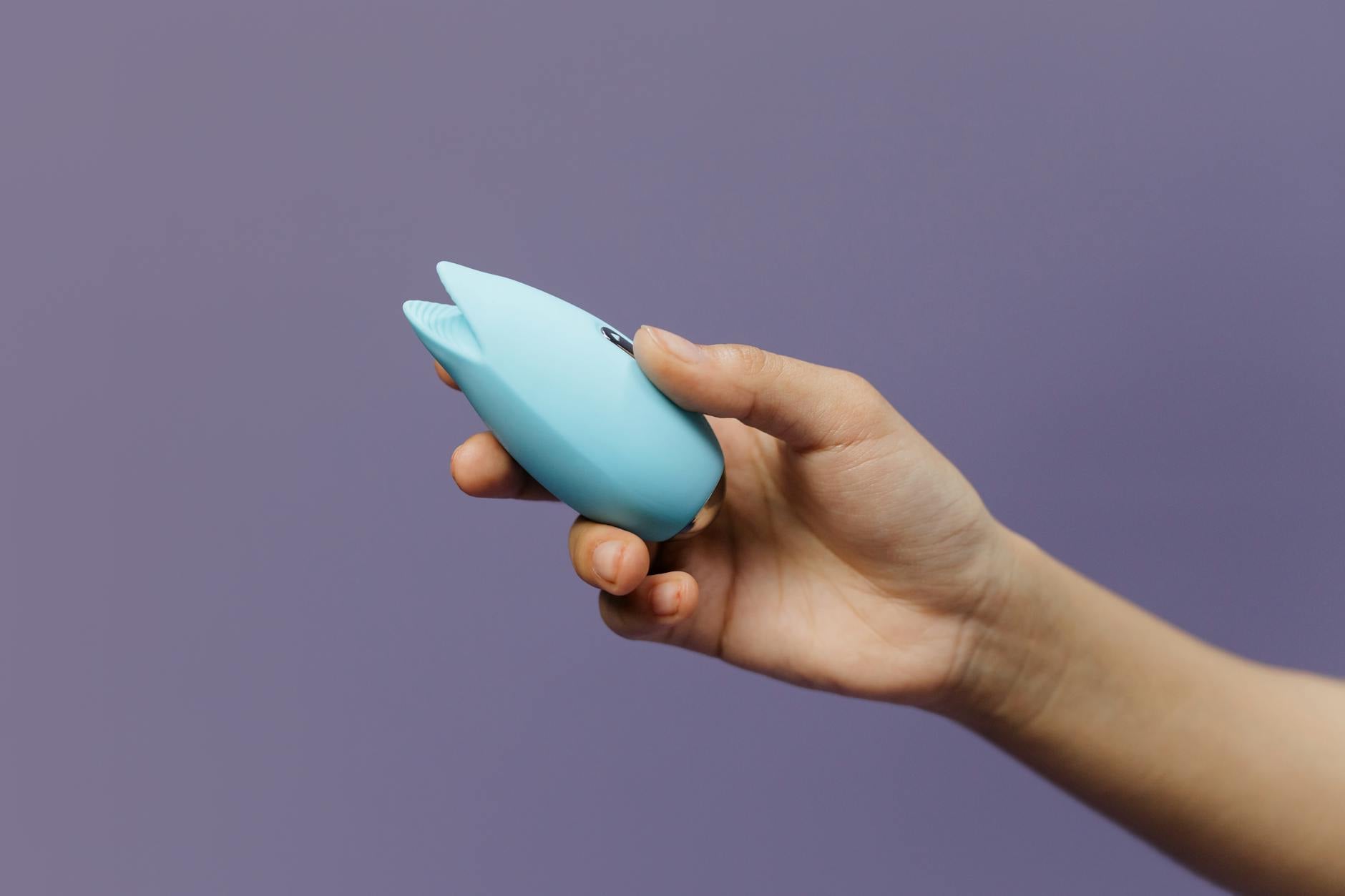 Vibrador de silicone azul sendo segurado na mão contra fundo roxo sólido, promovendo amor próprio e sexualidade